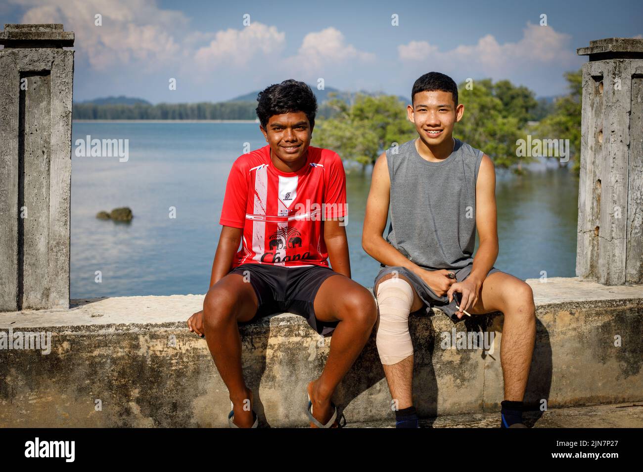 Dos adolescentes se sientan en un dique sonriendo, un niño con un cigarrillo, con el mar y el fondo tropical de la isla tailandesa Foto de stock