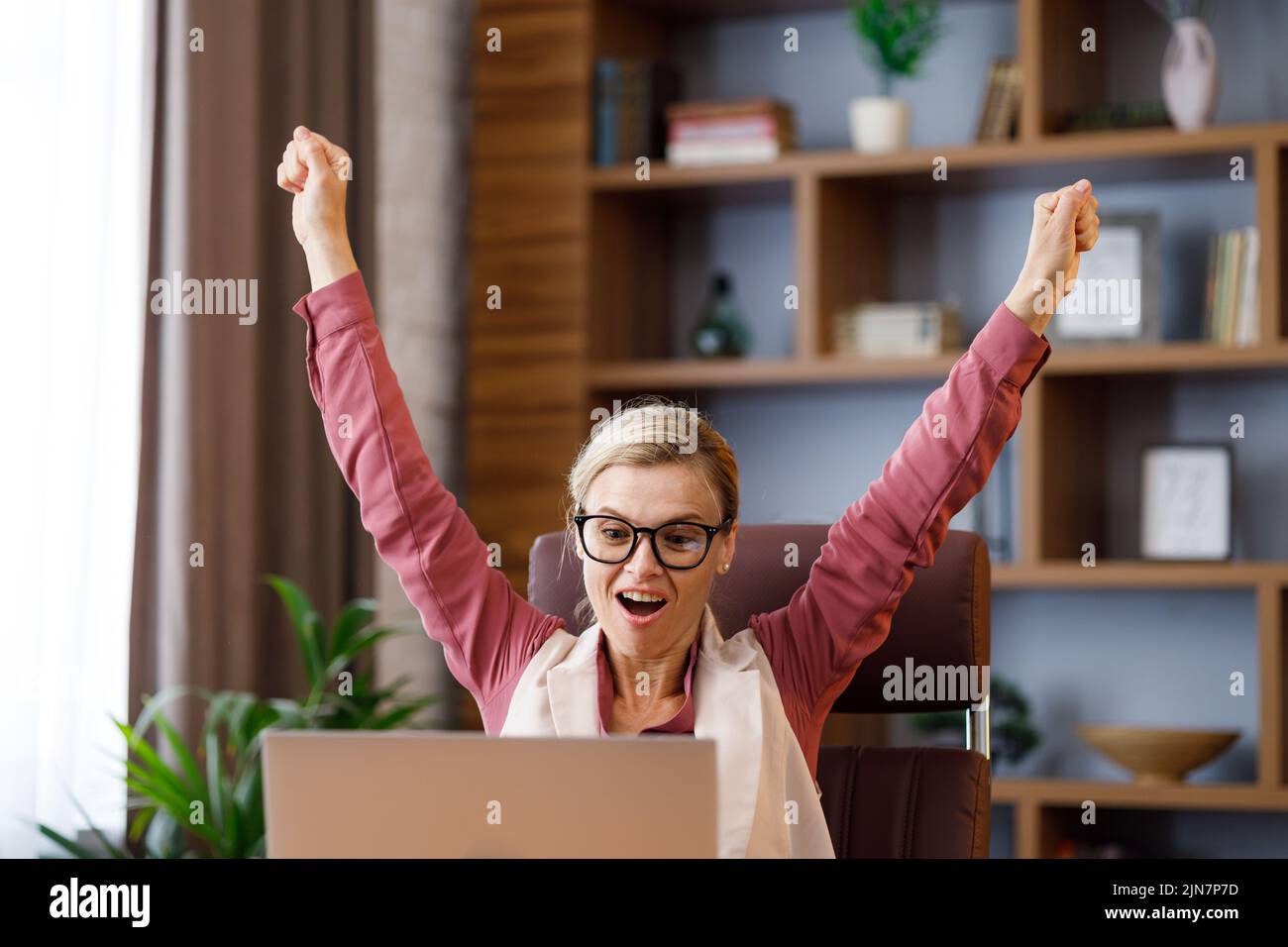 Feliz mujer de negocios sorprendida por las buenas noticias mientras mira el ordenador portátil sentado en la oficina. El gerente disfruta del éxito, celebra la victoria, consigue un buen trato Foto de stock
