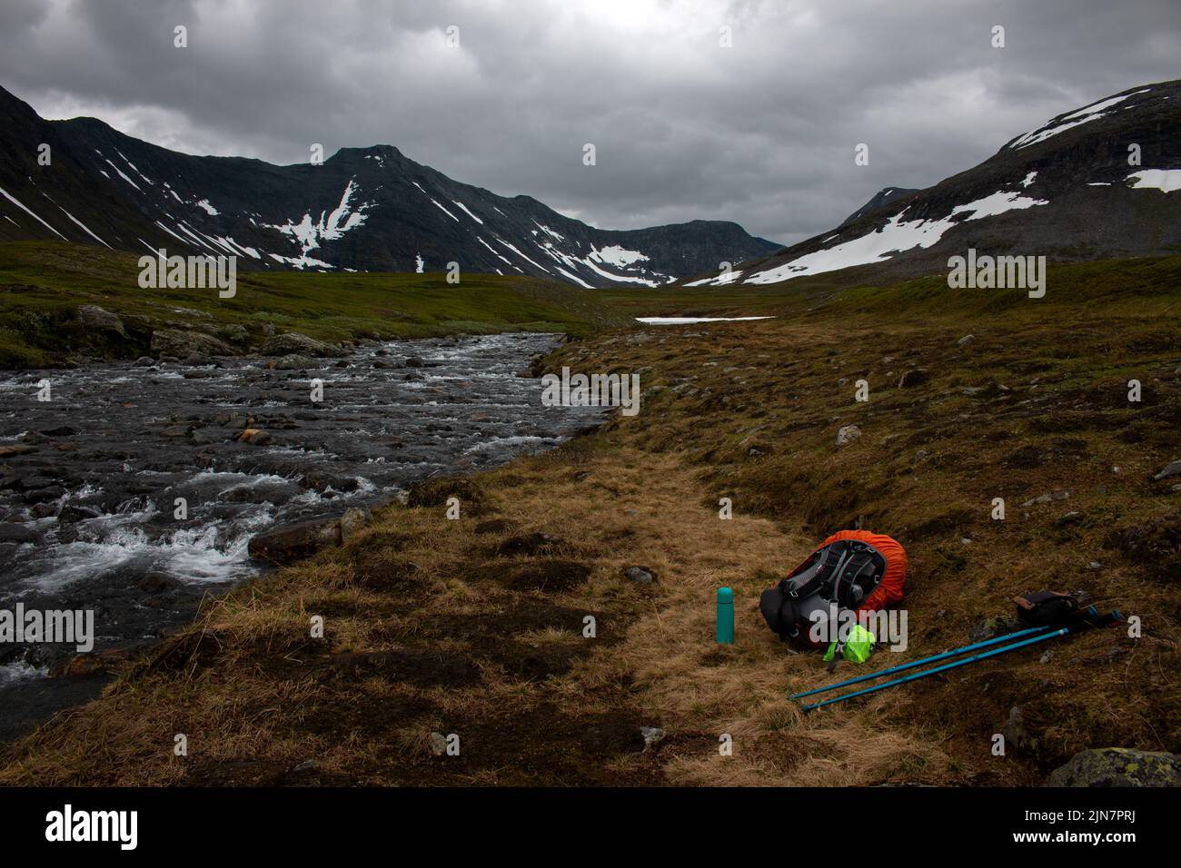 Una mochila, bastones de senderismo, y un termo junto a un arroyo que viene de la nieve que se derrite en la cima de las montañas Sylarna, Noruega Foto de stock