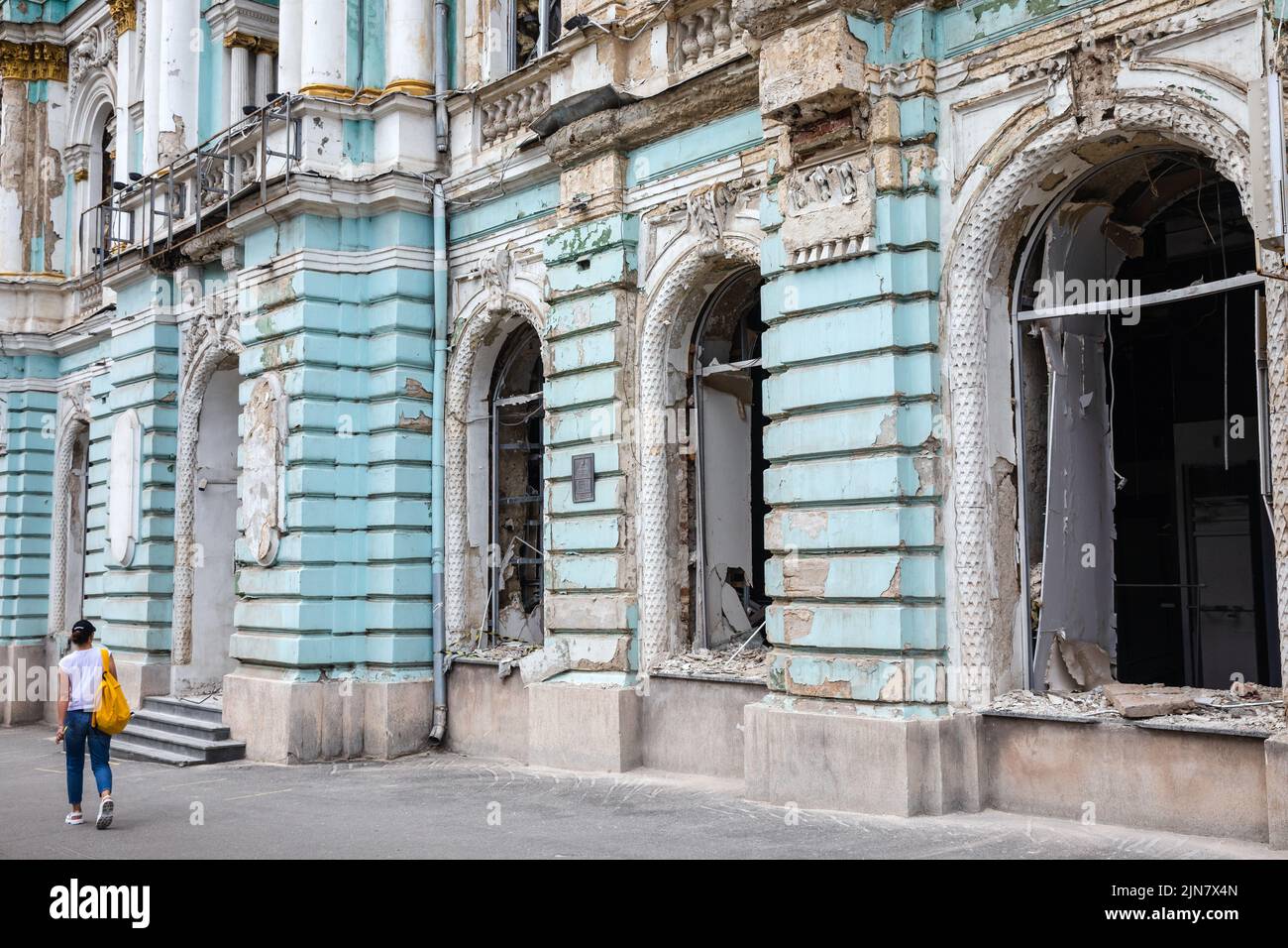 Una mujer joven camina por un edificio dañado con ventanas rotas y una fachada dañada debido al bombardeo ruso en Kharkiv. Foto de stock