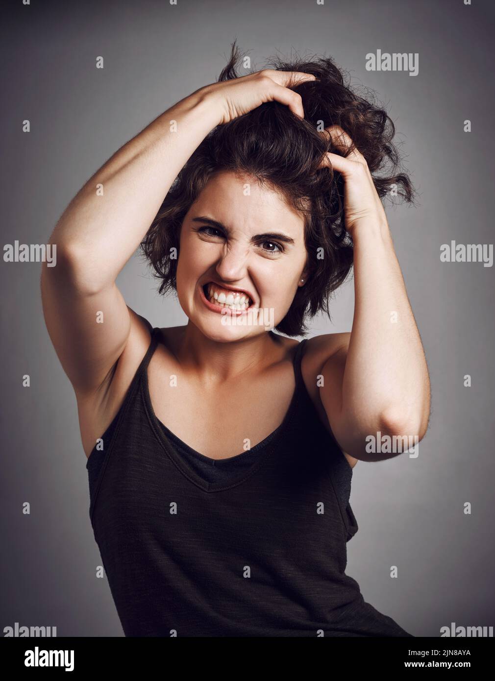 No puedo contener la rabia. Retrato de estudio de una atractiva mujer joven tirando de su pelo con rabia mientras se encuentra contra un fondo gris. Foto de stock
