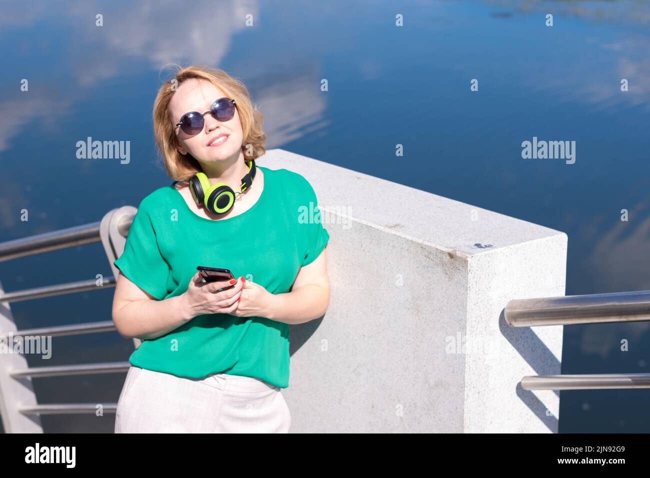 Una mujer sonriente con auriculares sosteniendo un teléfono en la mano escucha música de pie en la orilla del río. Una chica con auriculares y un teléfono en la parte trasera Foto de stock