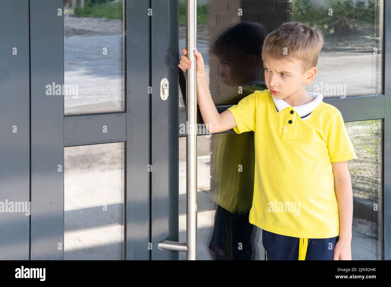 Un niño molesto sujetando el tirador de la puerta no puede abrir la puerta. El niño olvidó las llaves de la casa. El niño regresa a casa. Foto de stock