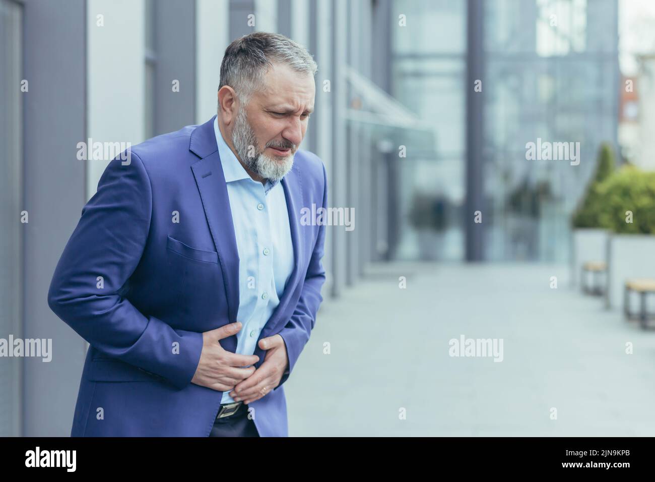 Hombre de negocios de pelo gris fuera del edificio de oficinas, hombre con traje de negocios enfermo tiene dolor de estómago. Foto de stock