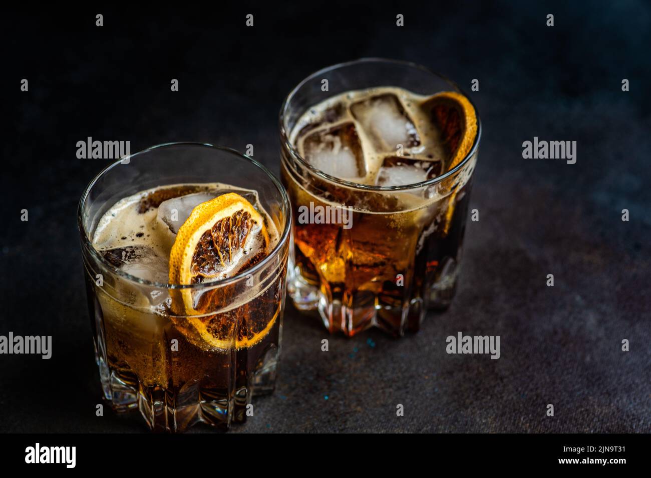 Cóctel de alcohol con coca-cola en el vaso con una rebanada de naranja y cubitos de hielo sobre una mesa de hormigón oscuro Foto de stock