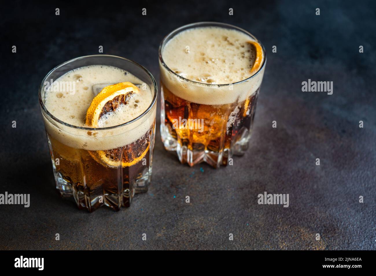 Cóctel de alcohol con coca-cola en el vaso con una rebanada de naranja y cubitos de hielo sobre una mesa de hormigón oscuro Foto de stock