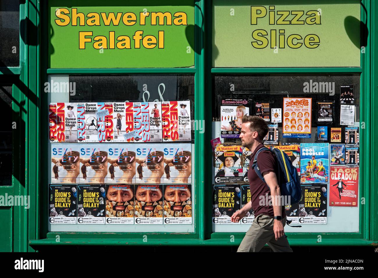 Un joven caminando por los carteles del Festival Fringe de Edimburgo pegados en la ventana de una pizzería para llevar. Foto de stock