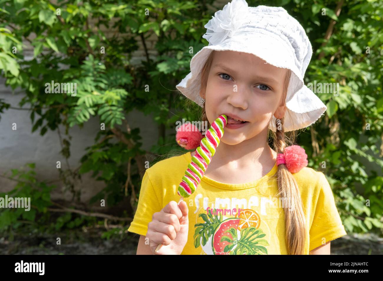 Una bonita chica con un sombrero blanco y un vestido amarillo lamiendo un colorido helado de caramelo sobre el fondo del follaje verde en el parque. horario de verano Foto de stock
