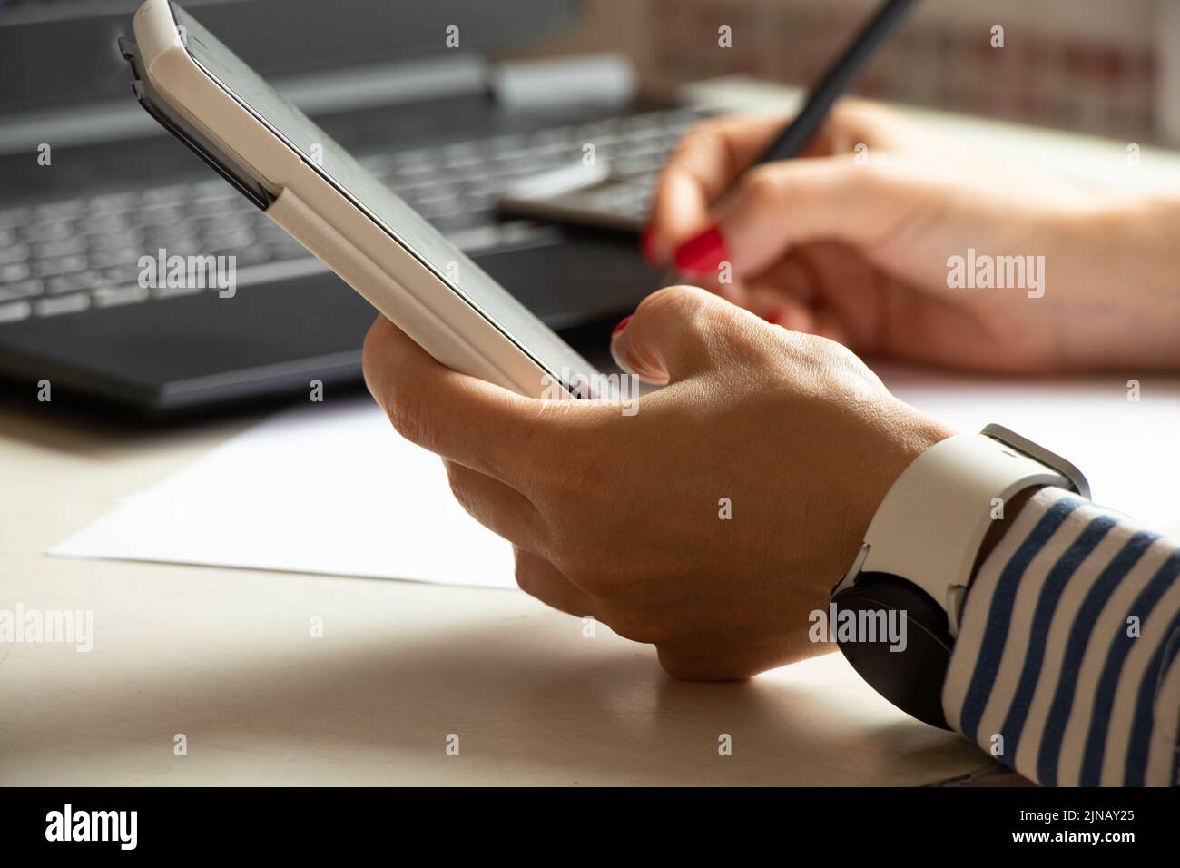 Una chica con camisas se sienta en una mesa con un teléfono en las manos, cerca de un portátil, cerca de una ventana en la oficina, trabajo en línea, móvil en sus manos Foto de stock