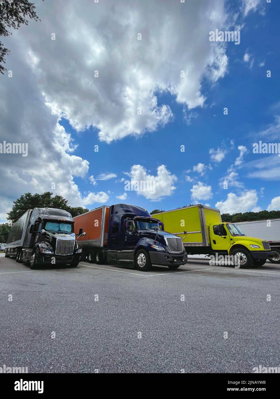 Tres camiones grandes y coloridos estacionados en asfalto en una parada de descanso de la carretera bajo un cielo azul brillante con nubes dramáticas Foto de stock