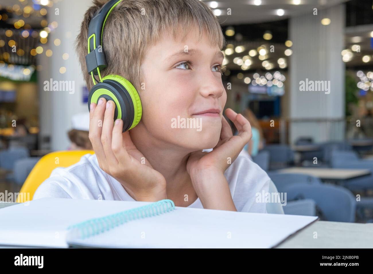 Un chico guapo escucha música con auriculares y escribe la tarea en un cuaderno en un café, bokeh, primer plano. Tecnología y música en la vida de los niños. Onlin Foto de stock