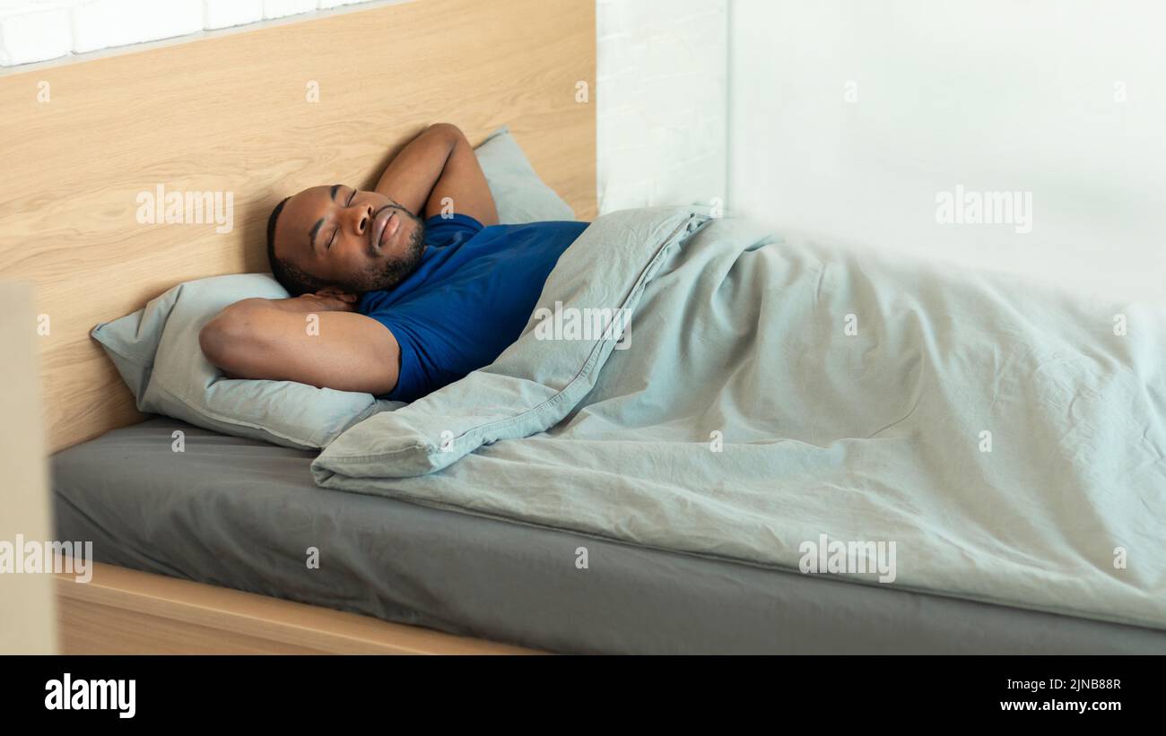 Hombre negro durmiendo en la cama sosteniendo las manos detrás de la cabeza Interior Foto de stock