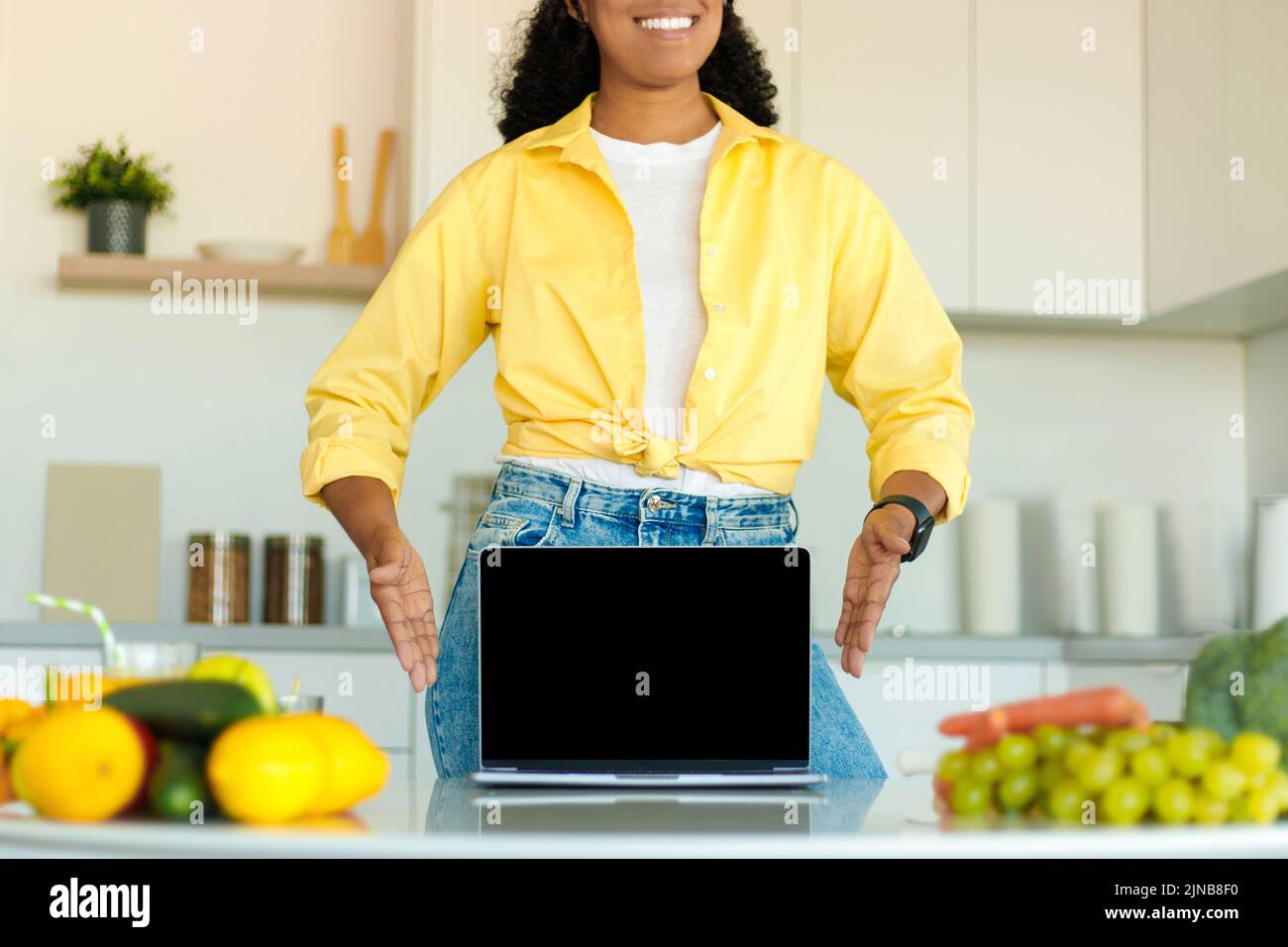 Mujer afroamericana mostrando portátil con pantalla en blanco en la mesa con verduras orgánicas en el interior de la cocina, maqueta Foto de stock