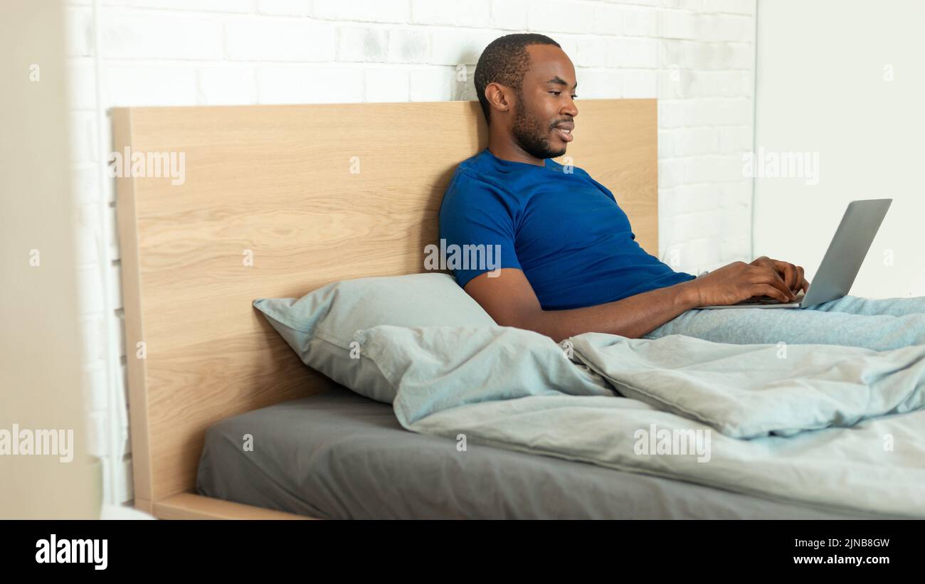 Hombre negro usando la escritura del ordenador portátil sentado en la cama en casa Foto de stock