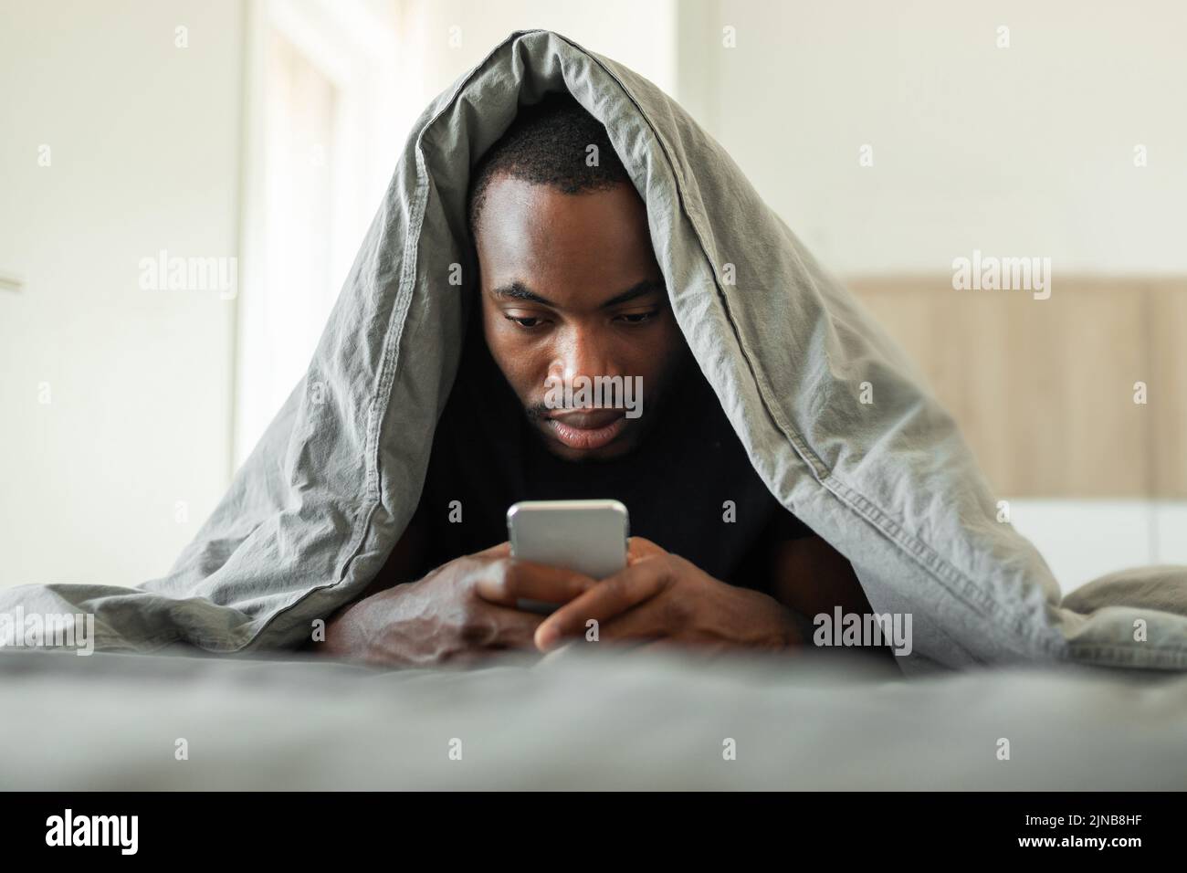 Hombre Afroamericano soñoliento que utiliza el teléfono celular que se encuentra en la cama interior Foto de stock