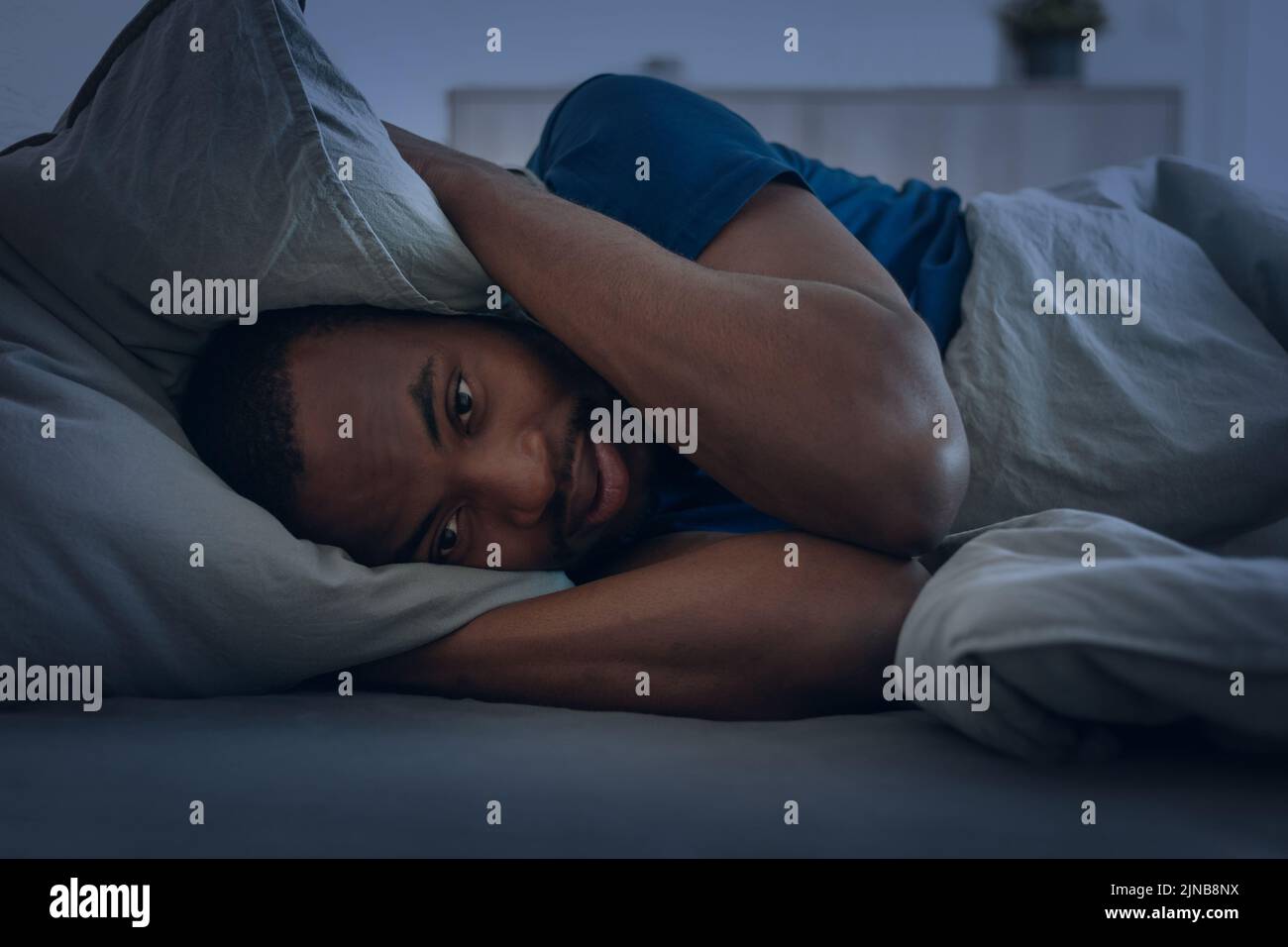 Hombre negro que tiene insomnio cubriendo las orejas con almohada en el dormitorio Foto de stock