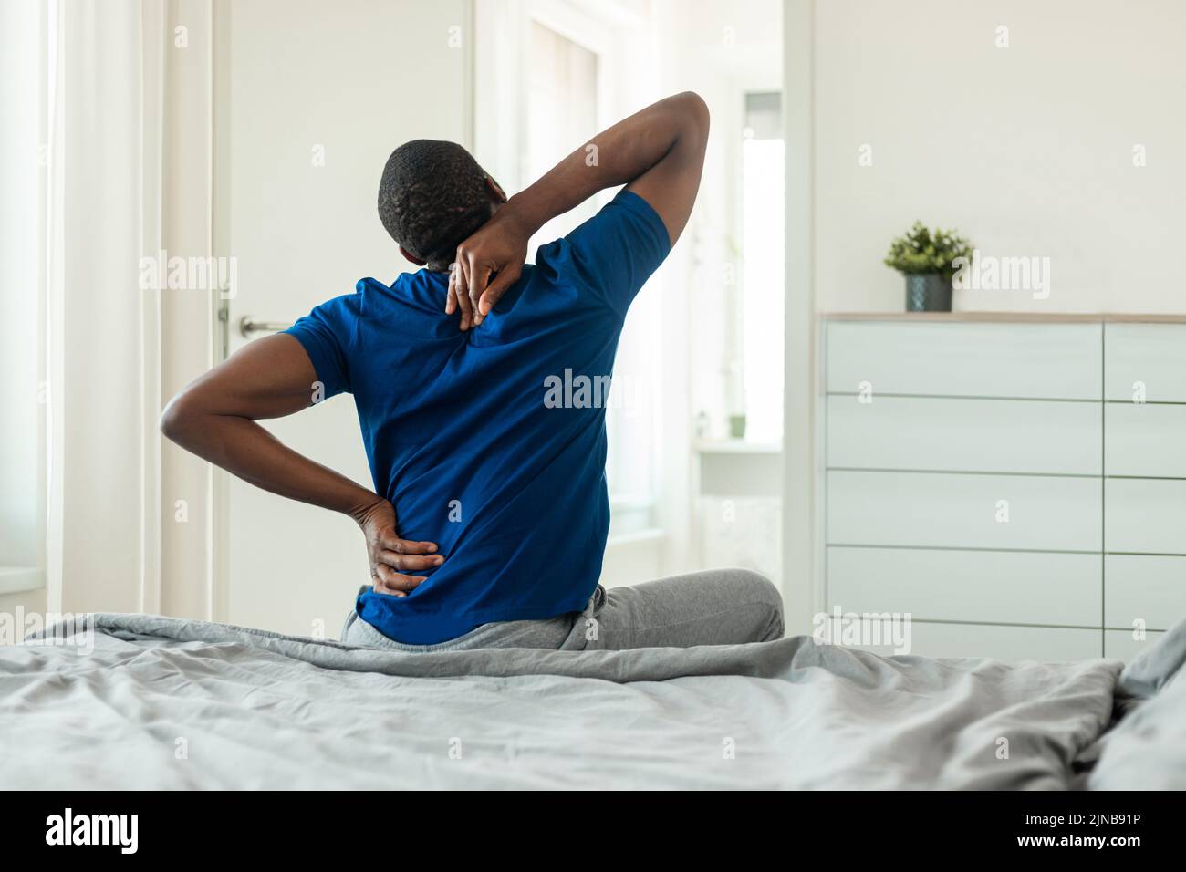 Hombre africano tocando el cuello dolorido y la columna sentado en el dormitorio Foto de stock