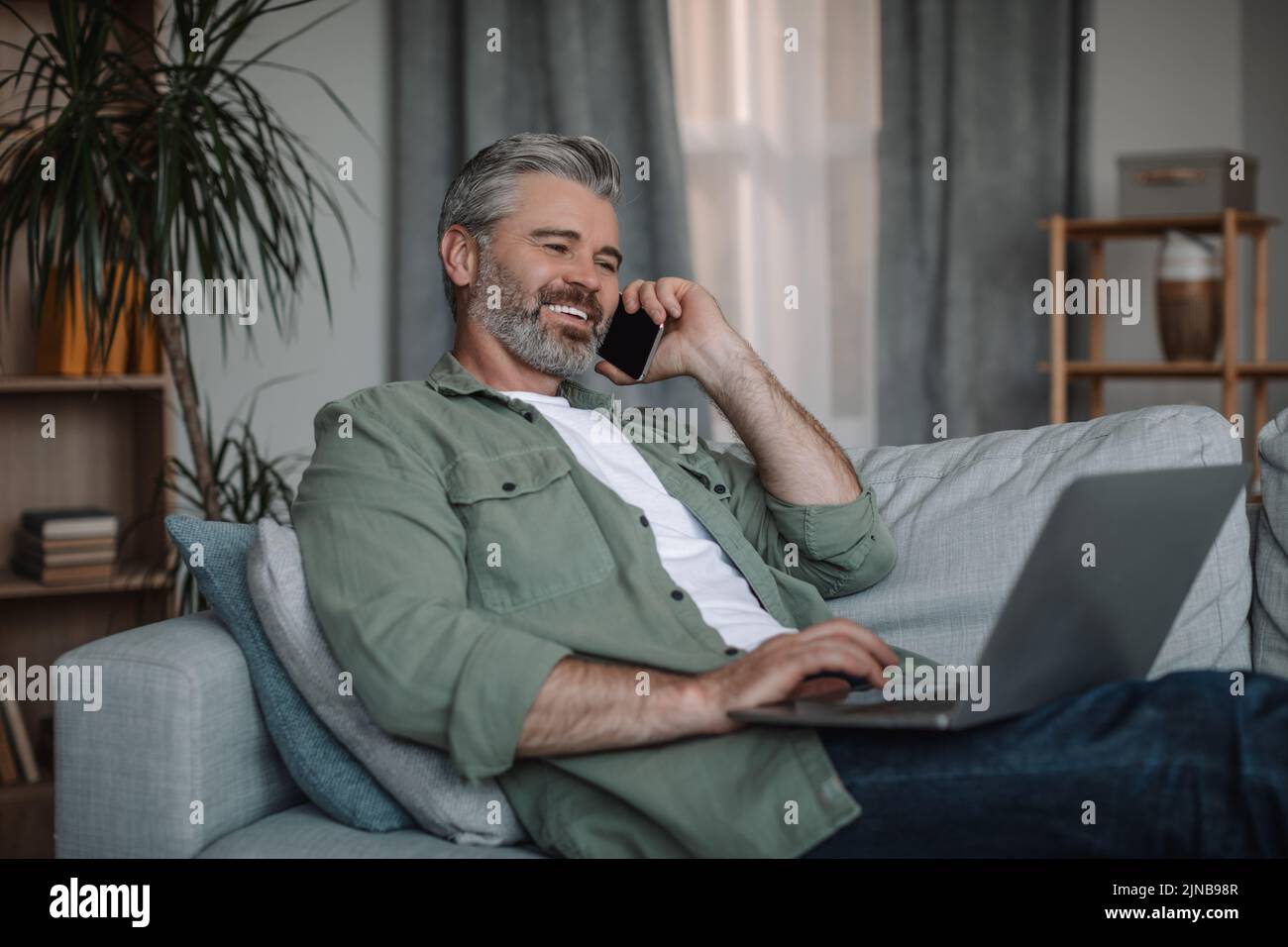 Alegre hombre europeo maduro con llamada para la barba por teléfono, trabaja en el portátil, se sienta en el sofá en el interior de la sala de estar Foto de stock