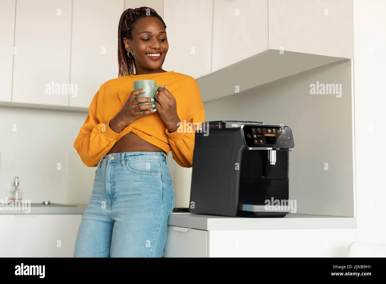 Feliz mujer afroamericana disfrutando de café aromático fresco cerca de la máquina moderna en el interior de la cocina Foto de stock