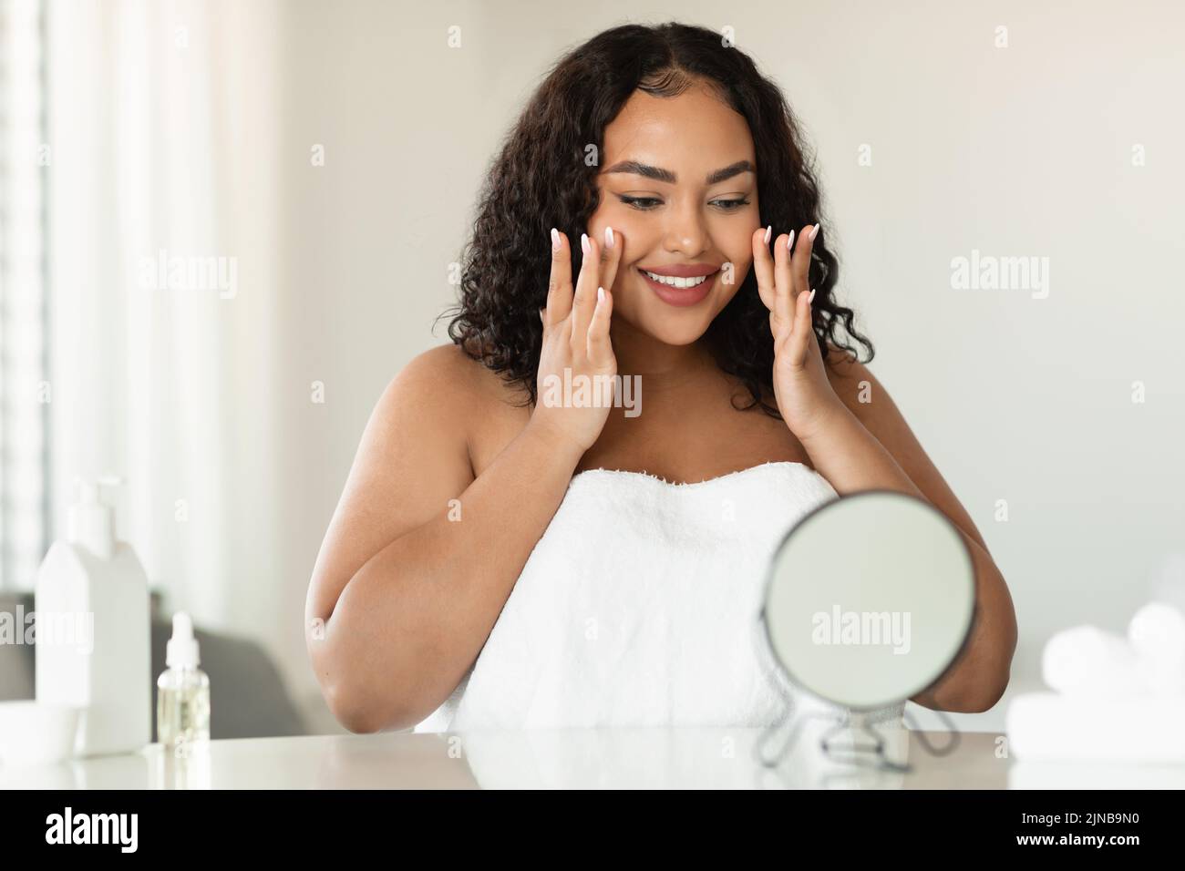 Concepto de estiramiento facial y cuidado de la piel. Joven mujer negra más tamaño masajeando su rostro y mirando el espejo Foto de stock