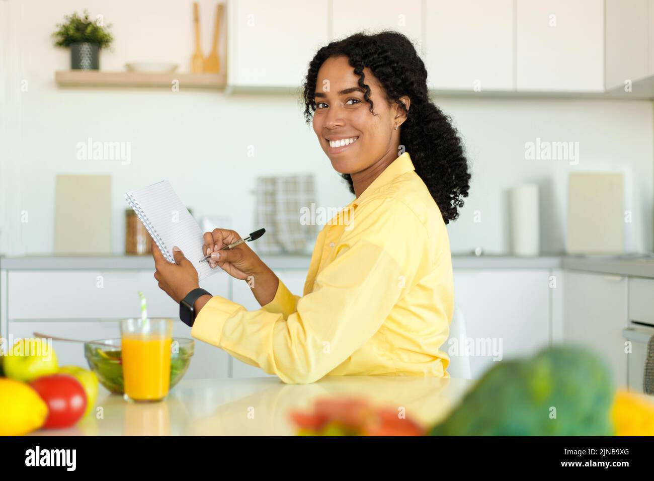 Concepto de plan de dieta. Feliz dama negra escribiendo dieta diaria ración o menú en el cuaderno, sentado en la mesa de la cocina Foto de stock