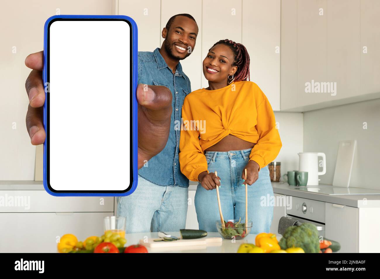 Emocionado negro cónyuges preparar ensalada con verduras orgánicas juntos, el hombre muestra gran smartphone con pantalla en blanco Foto de stock