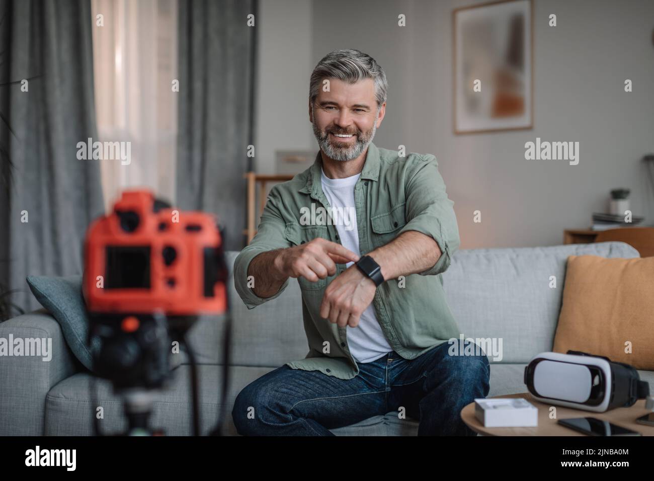 Alegre hombre europeo retirado con barba hace una visión general de los dispositivos y muestra el reloj inteligente Foto de stock