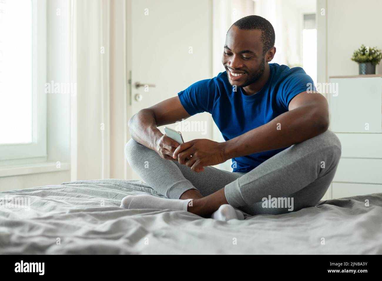 Hombre negro usando mensajes de texto de smartphone sentado en el dormitorio de casa Foto de stock