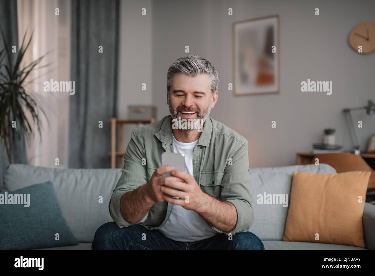 Alegre hombre europeo anciano con barba, charlando con smartphone, viendo vídeo, jugando Foto de stock