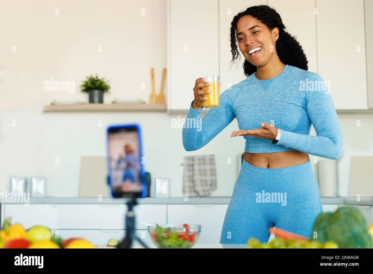 FIT Black Woman grabando vídeo sobre alimentación saludable, mostrando un vaso de zumo fresco en la cámara del smartphone en la cocina Foto de stock