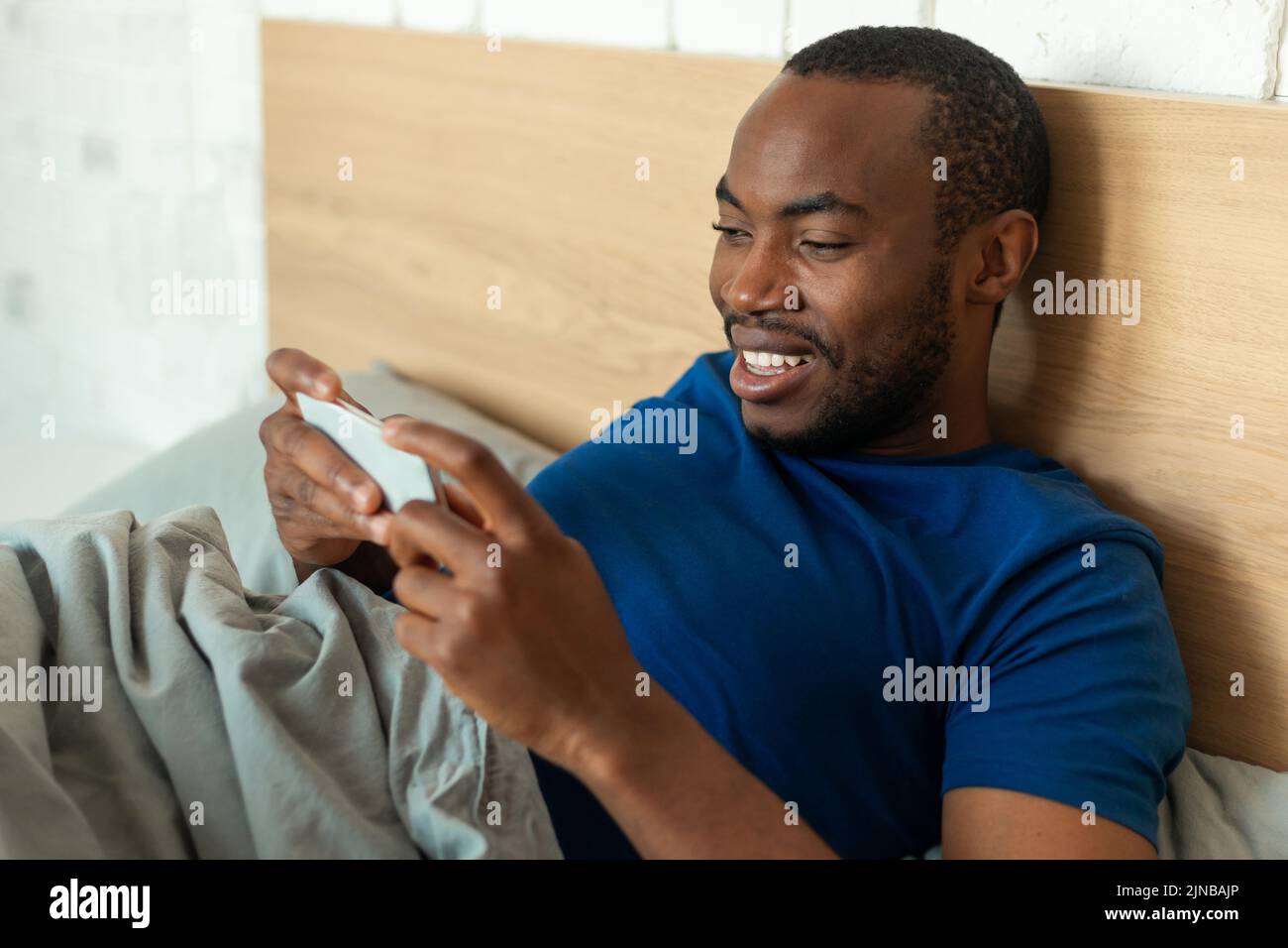 Hombre africano usando el teléfono jugando juego acostado en la cama interior Foto de stock