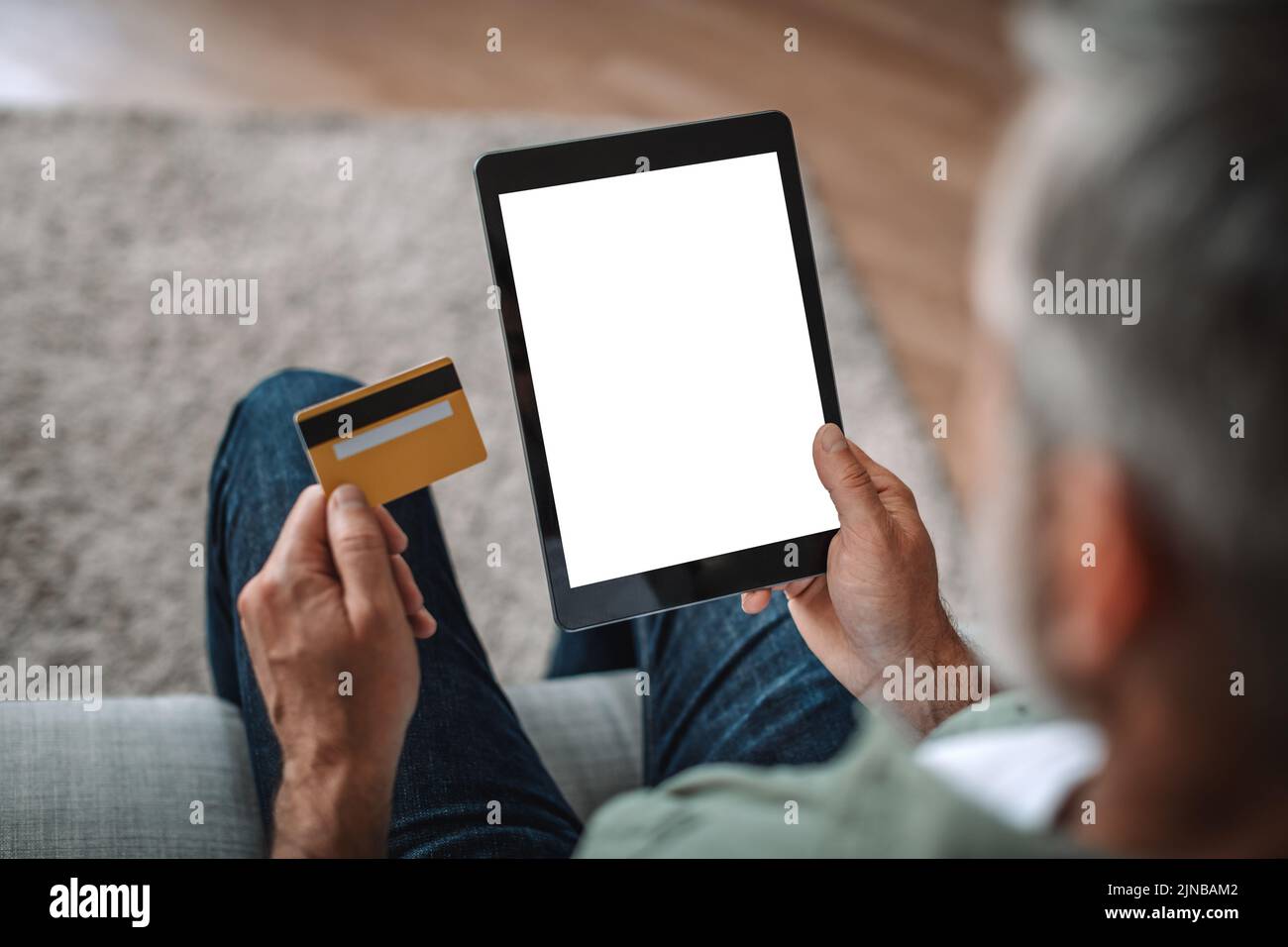 El hombre europeo jubilado con barba utiliza tarjeta de crédito y tablet con pantalla vacía, comprando en línea en el salón Foto de stock