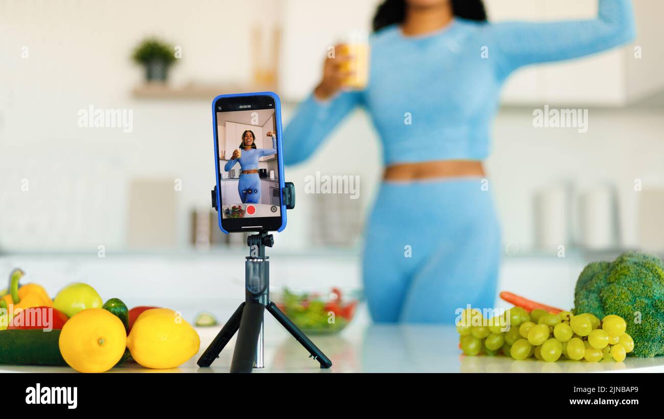 Excitada mujer negra de ajuste grabando nuevo vídeo para su blog de deporte y comida en el smartphone, de pie en la cocina Foto de stock