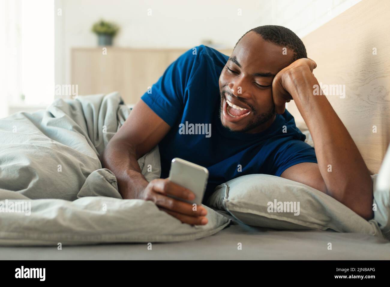 Hombre negro alegre con un smartphone tumbado en la cama en la habitación Foto de stock
