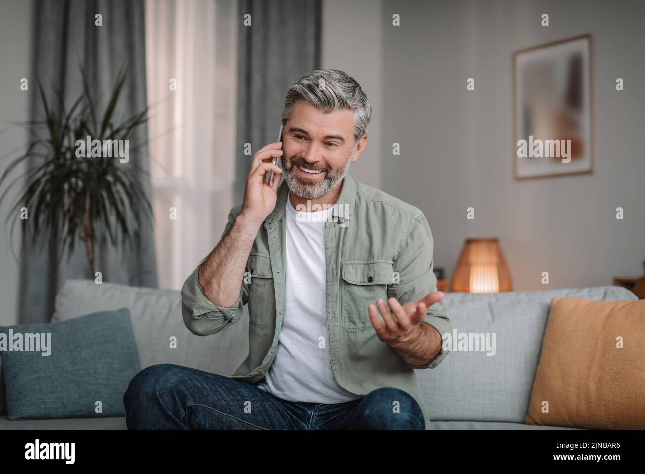 Sonriente hombre europeo jubilado con llamadas para la barba mediante smartphone, mando a distancia en el interior del salón Foto de stock