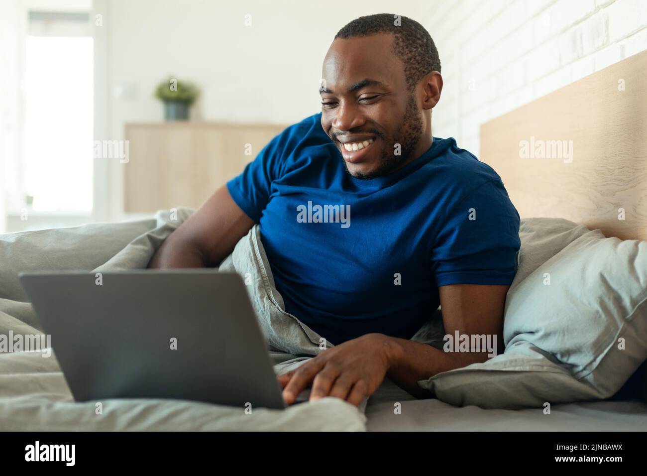 Hombre africano que utiliza el ordenador portátil Trabajando en línea acostado en la cama interior Foto de stock