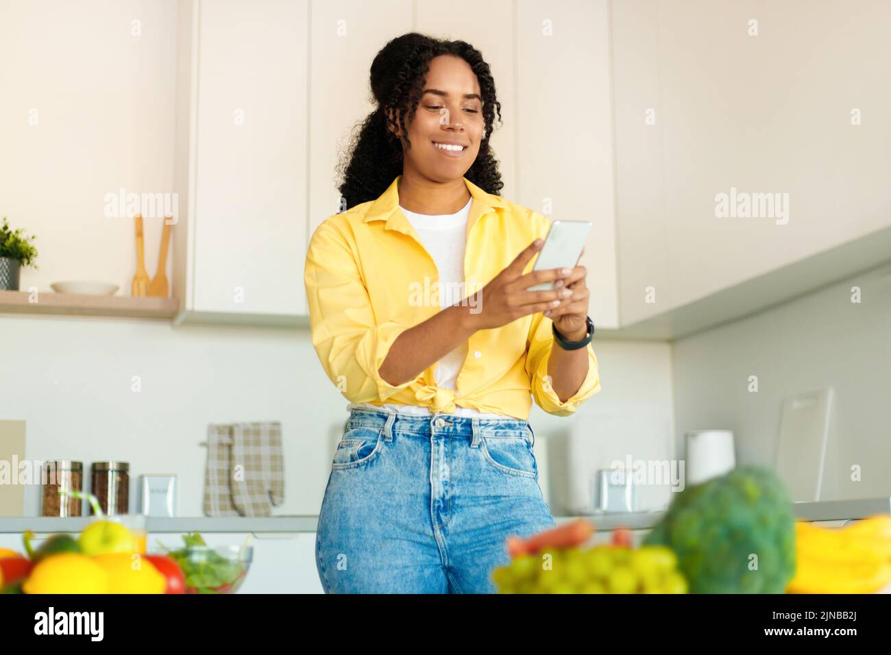 Pedidos en línea, alimentos saludables. Mujer negra feliz con smartphone en el interior de la cocina, espacio de copia gratuito Foto de stock