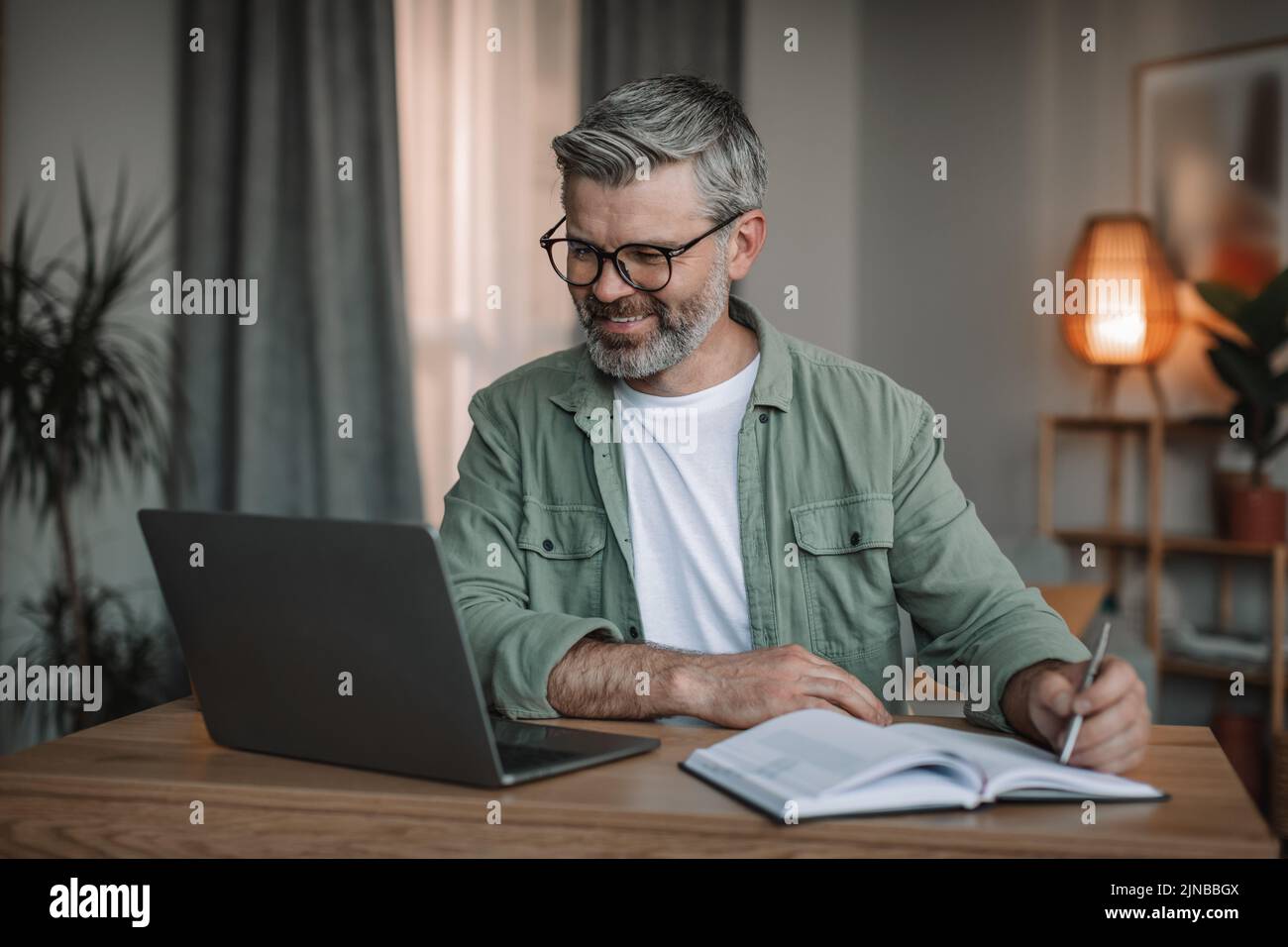 Alegre hombre europeo senior con barba en gafas, mira el vídeo en el ordenador, toma notas en la mesa de la habitación Foto de stock