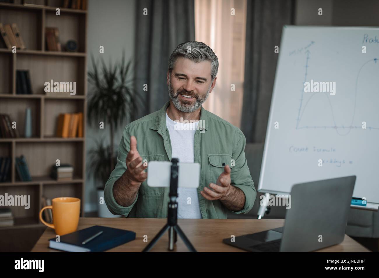 Alegre hombre europeo retirado con profesor de barba que da una lección de vídeo con un smartphone en el interior de la habitación Foto de stock