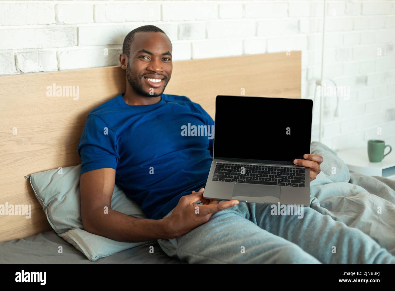 Hombre afroamericano sosteniendo ordenador portátil con pantalla vacía en el dormitorio Foto de stock