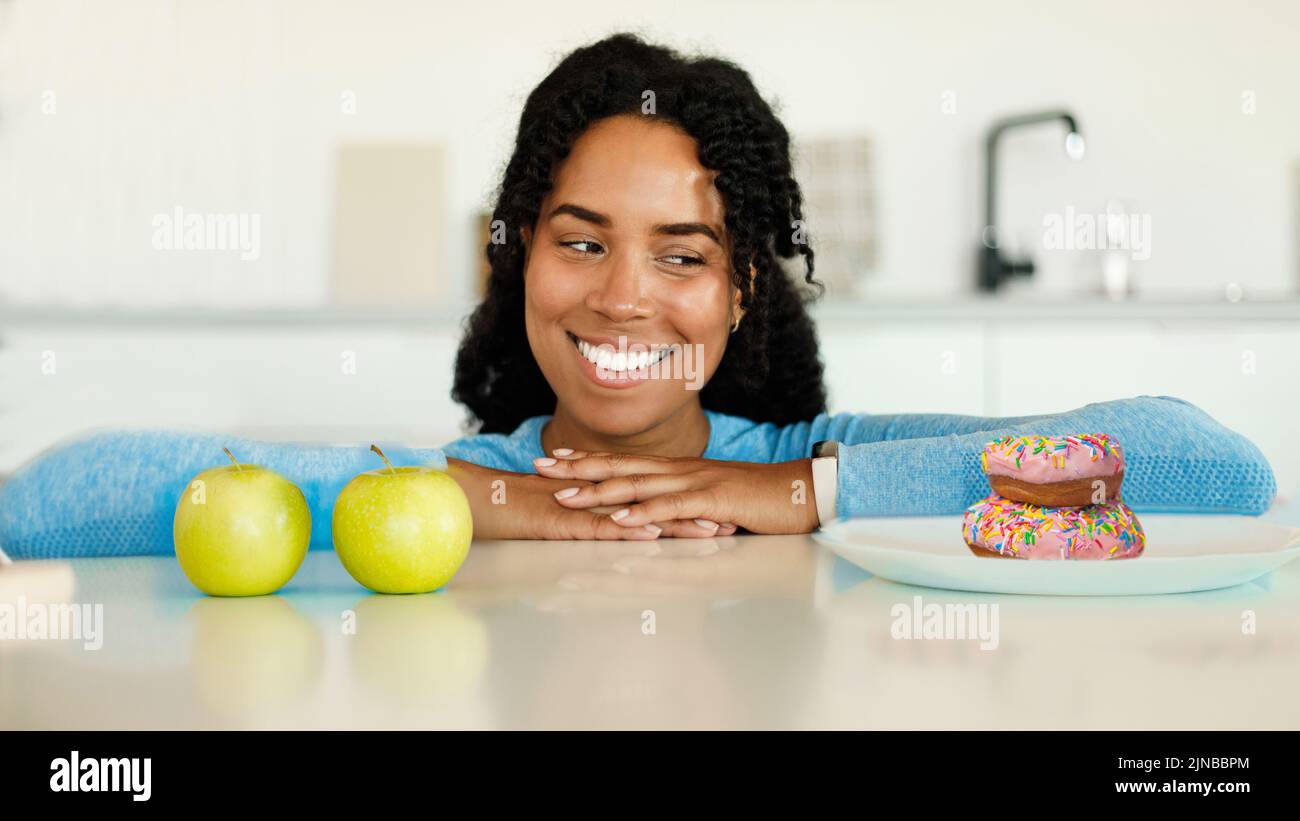 Dieta saludable vs no saludable. Mujer negra mirando donuts y manzanas, haciendo elección de su comida Foto de stock