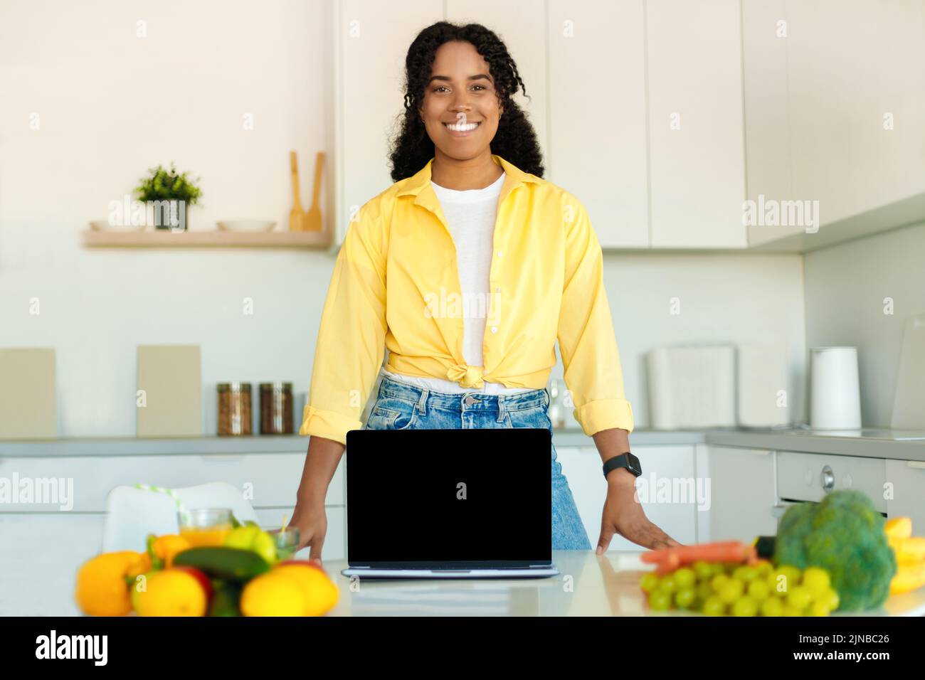 Recetas de cocina en línea. Feliz dama negra mostrando el ordenador portátil con pantalla en blanco en la cocina moderna, maqueta, espacio de copia Foto de stock