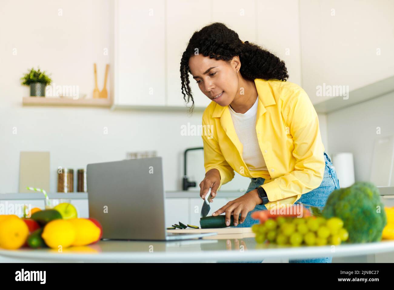 Mujer afroamericana cocinando ensalada orgánica fresca en casa en la cocina moderna, mirando el ordenador portátil, probando nueva receta Foto de stock