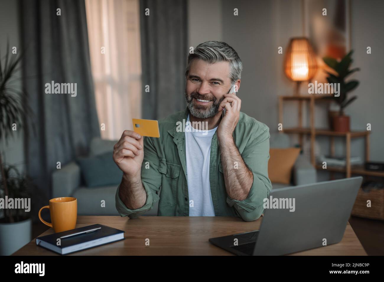 Feliz retirado hombre caucásico con barba llamadas por teléfono inteligente, utiliza tarjeta de crédito y pc para hacer compras en línea Foto de stock