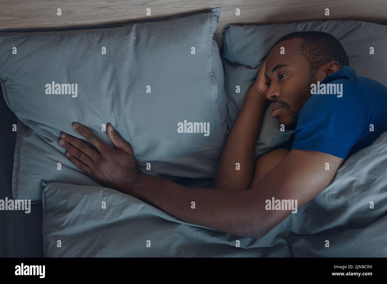 Infeliz hombre negro tocando almohada acostada en la cama interior Foto de stock