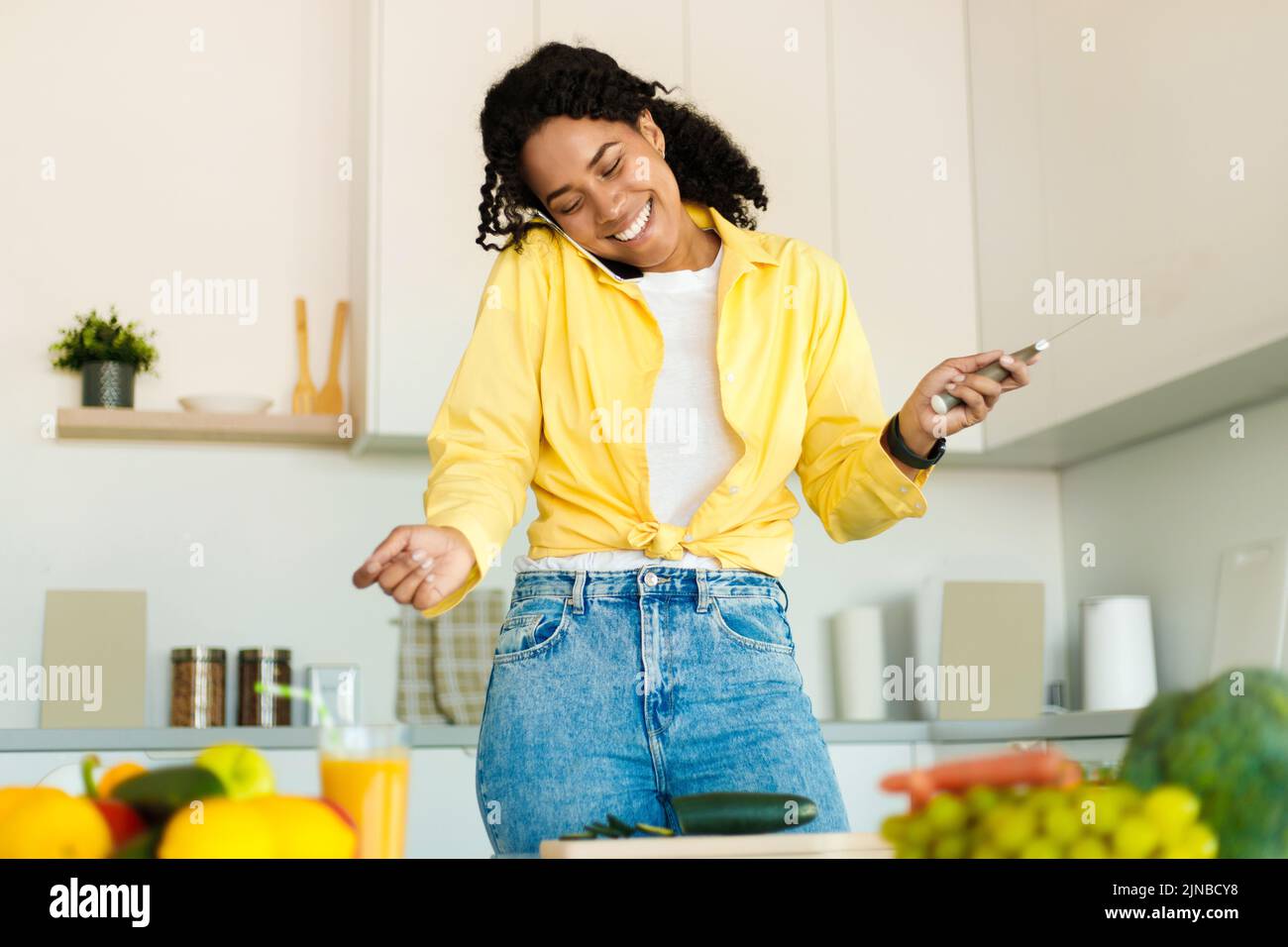 Alegre joven mujer negra hablando en el teléfono celular y sosteniendo el cuchillo en la mano, cortando pepino, preparando ensalada fresca Foto de stock