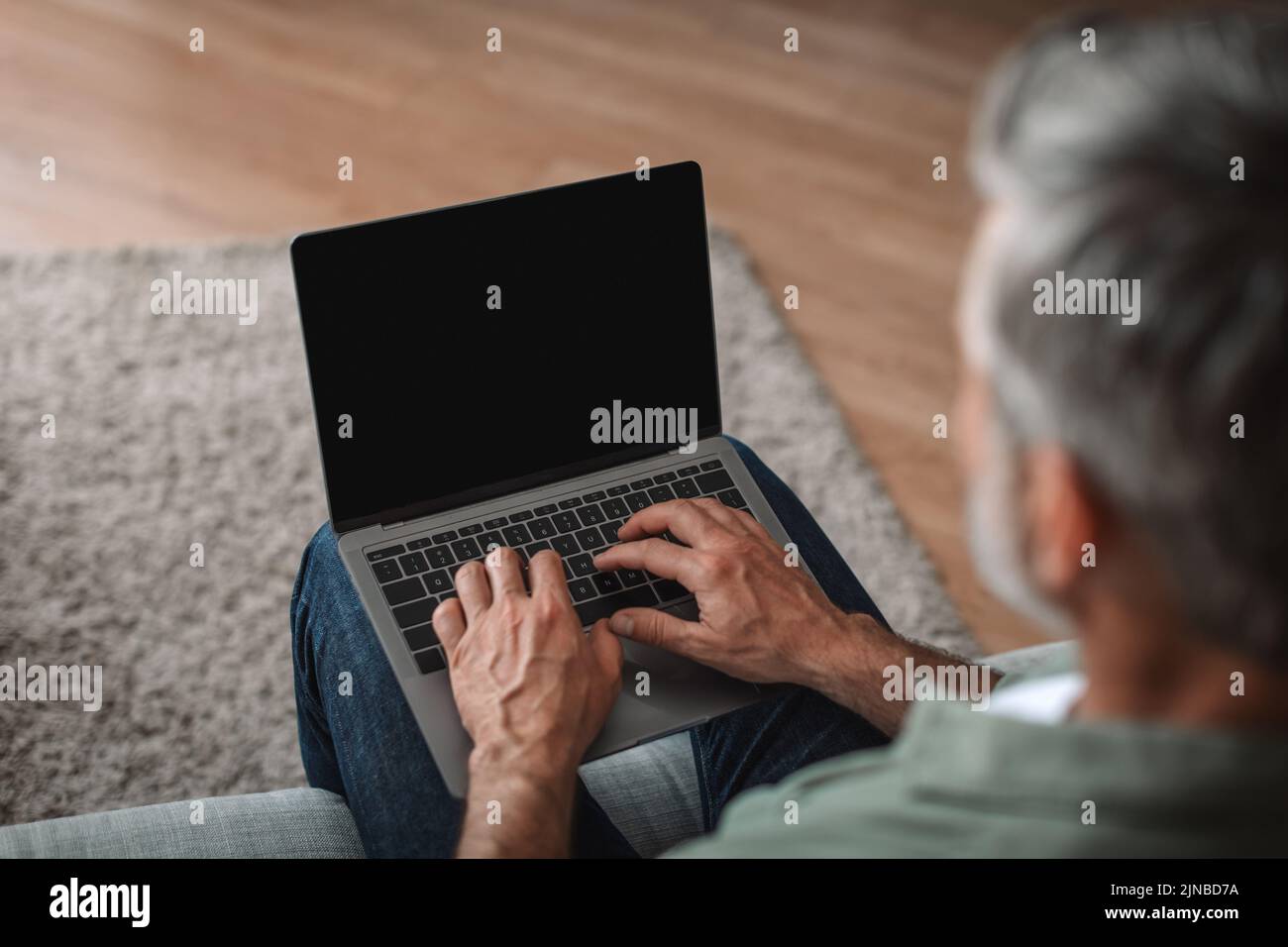 Hombre europeo mayor charlando en un portátil con pantalla en blanco, viendo vídeo en el interior del salón Foto de stock