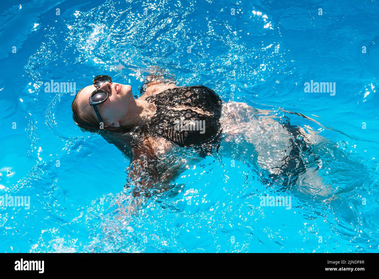 Mujer hermosa en una piscina. Chica en traje de baño negro y gafas de sol nada en su espalda en agua clara en la piscina Foto de stock