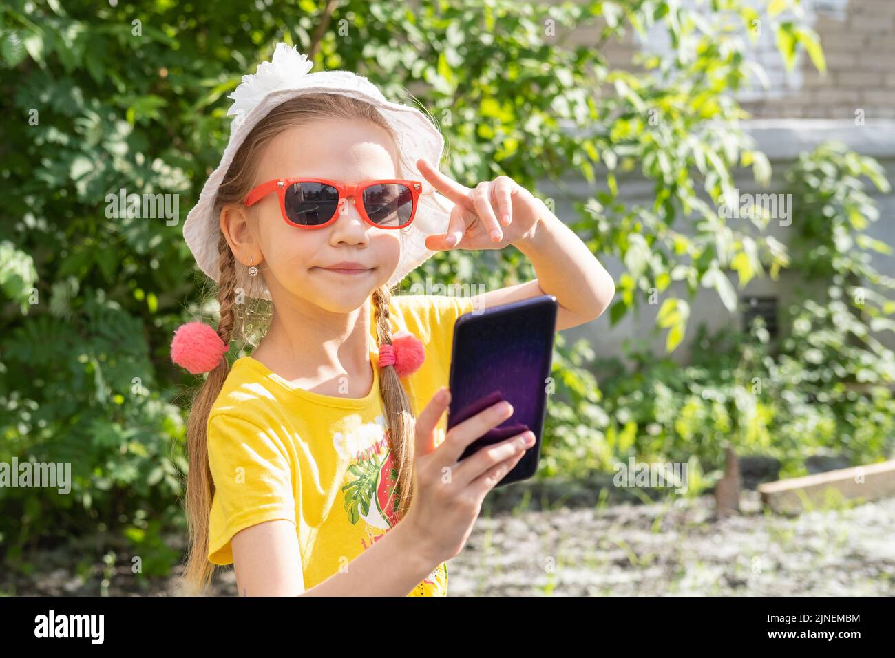 Retrato de una niña haciendo un selfie con un smartphone en el parque de verano. Una niña alegre en un vestido amarillo, sombrero blanco y gafas de sol lleva un Foto de stock