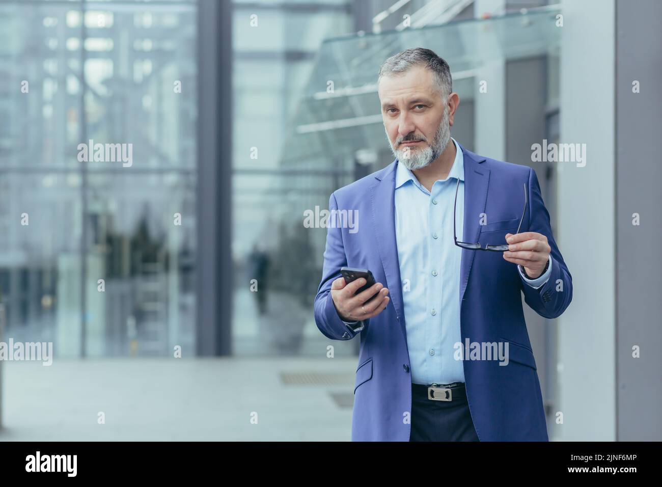 Retrato de un financiero senior de pelo gris, un hombre fuera del edificio de oficinas sosteniendo el teléfono y mirando la cámara, un hombre de negocios caminando Foto de stock